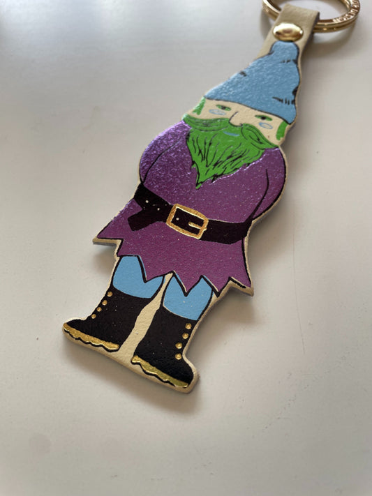 A gnome-shaped keychain charm with a colorful hand-painted design, featuring a metallic purple coat, green beard, blue hat, and black boots, attached to a gold-colored split ring on a light gray background.