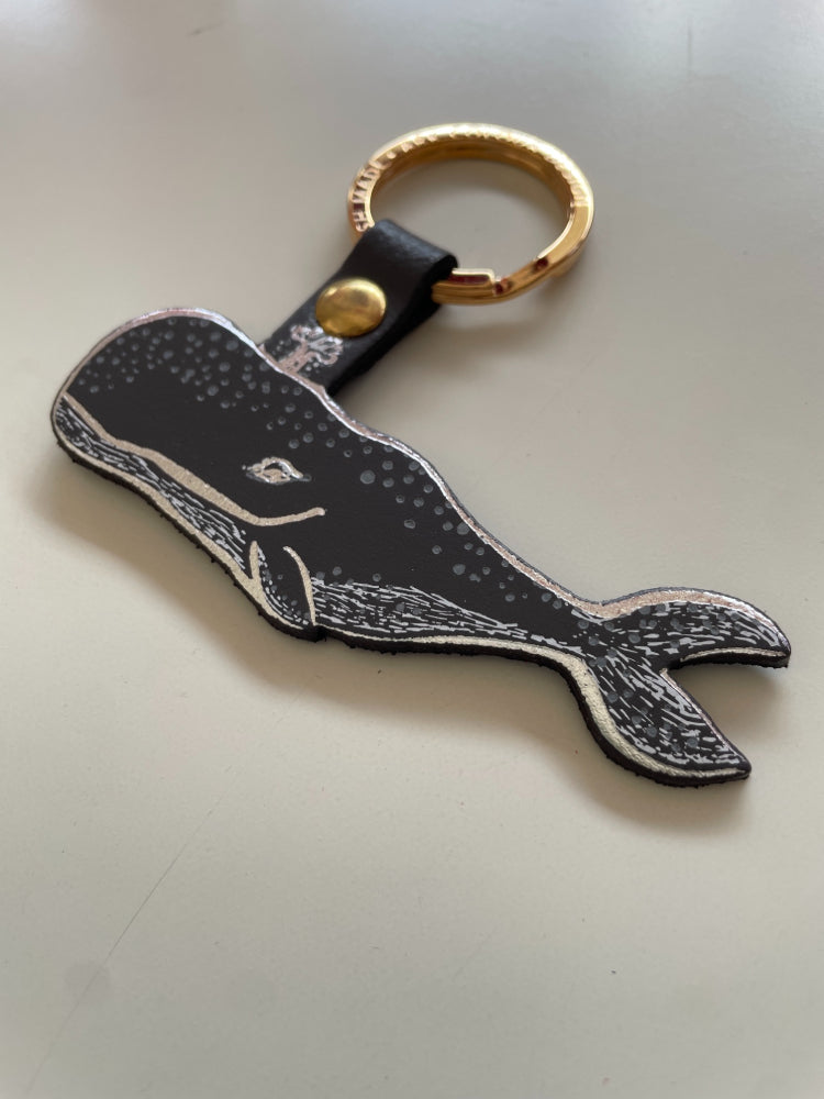 A whale-shaped keychain with a dark grey and white spotted design, attached to a dark grey strap with a gold snap and a gold split ring, shown on a white surface.