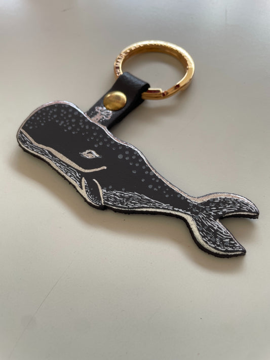 A whale-shaped keychain with a dark grey and white spotted design, attached to a dark grey strap with a gold snap and a gold split ring, shown on a white surface.