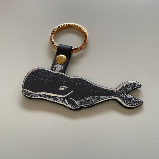A whale-shaped keychain with a dark grey  and white textured body, a gold-tone split ring, and a dark grey strap with a gold snap, photographed on a plain light background.