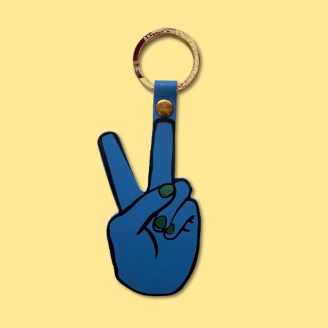 A blue leather keychain shaped like a hand making a peace sign with metallic green fingernails, with a gold-tone metal key ring attached at the top, shown on a solid pale yellow background.
