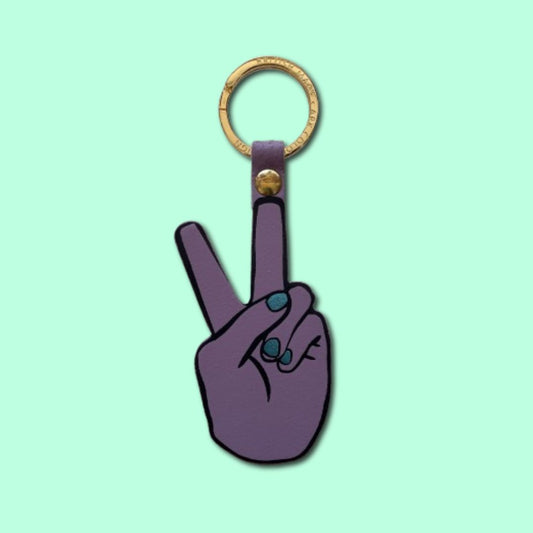 A purple leather keychain shaped like a hand making a peace sign with metallic blue fingernails, with a gold-tone metal key ring and a small gold rivet, shown on a solid mint green background.