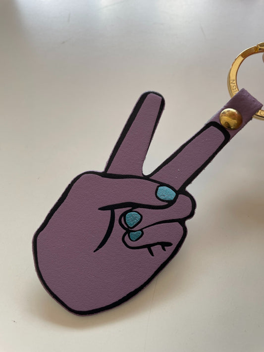 A purple leather keychain shaped like a hand making a peace sign and metallic blue fingernails, with turquoise nail accents and a gold-tone split ring.