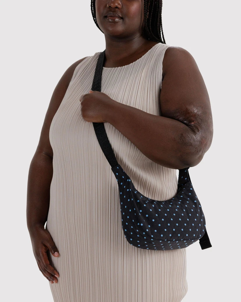 A model wearing a beige pleated dress carries a black shoulder bag with a light blue polka-dot pattern and black strap against a light gray background.