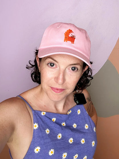 Person wearing a pink cap with an orange cat embroidered and a blue sleeveless top with white patterns against a plain background