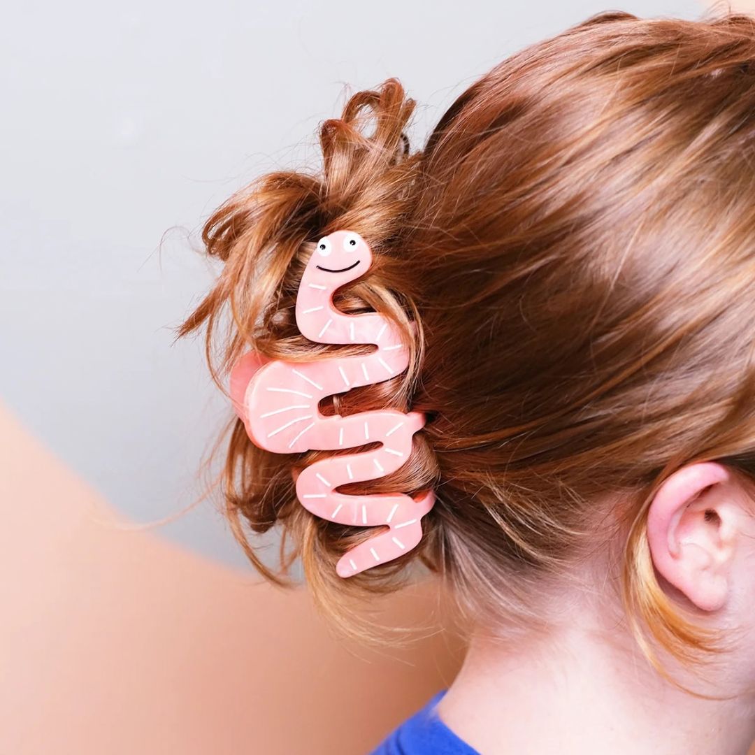 A pink worm-shaped hair claw clip holding a messy bun in a person's light red-brown hair, shown from a side-back angle against a soft gray background.