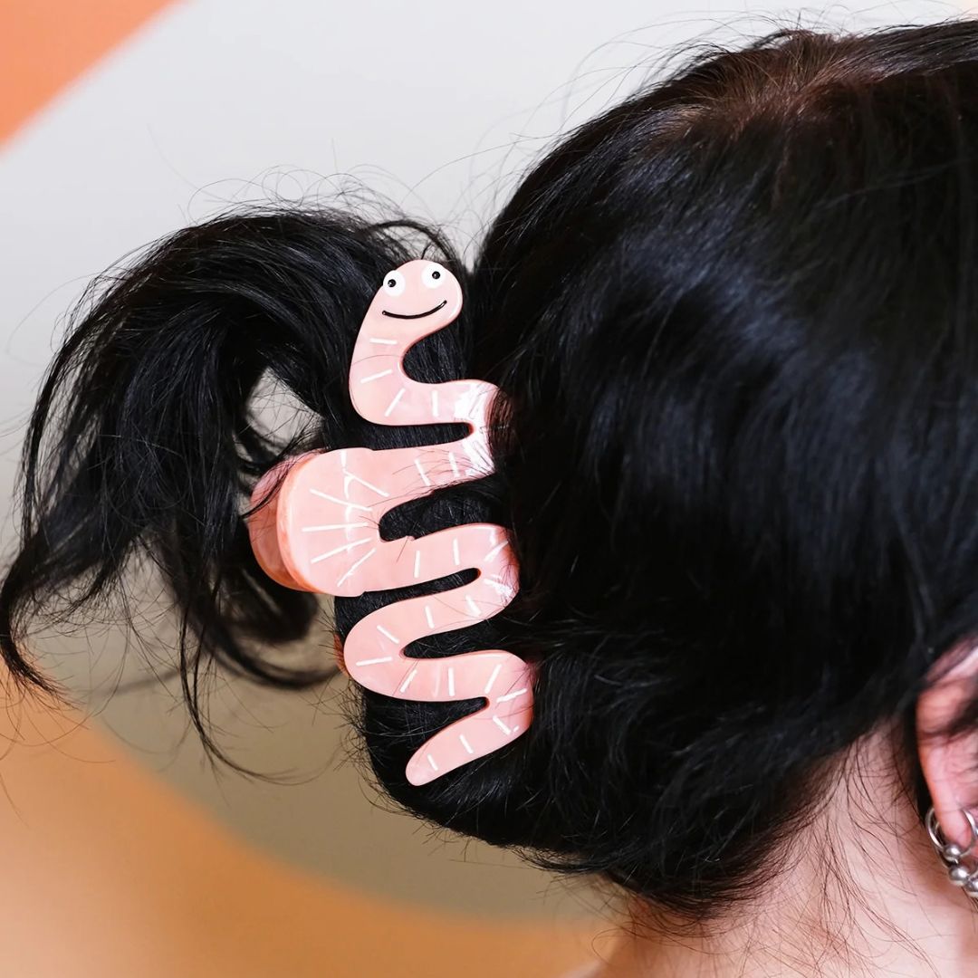 A pink wavy hair claw clip shaped like a smiling worm, holding up a section of black hair on a person's head.