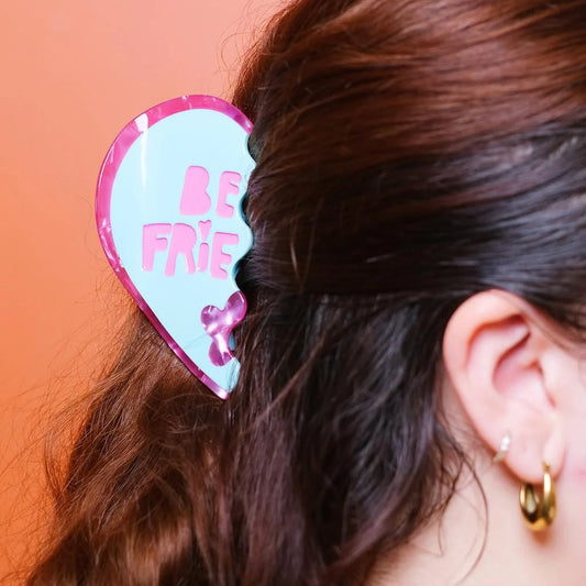 Heart-shaped hair clip with 'Be Friends' text worn in hair against an orange background