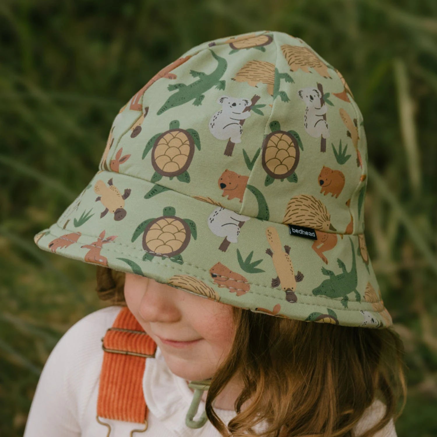 Side view of toddler wearing an Australian animal patterned bucket hat against a blurred green background