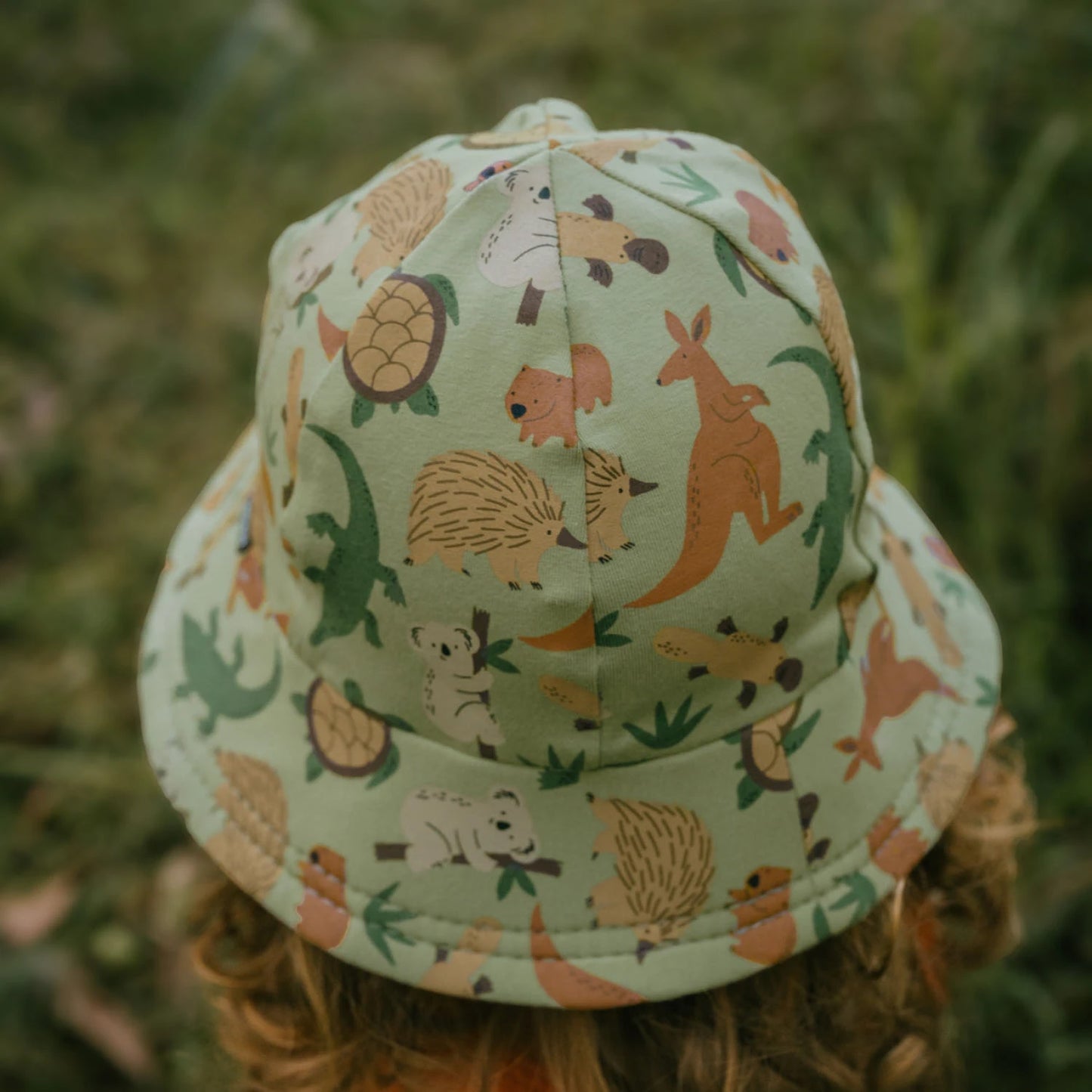 Back view of a Toddler wearing an Australian animal patterned bucket hat in a grassy setting