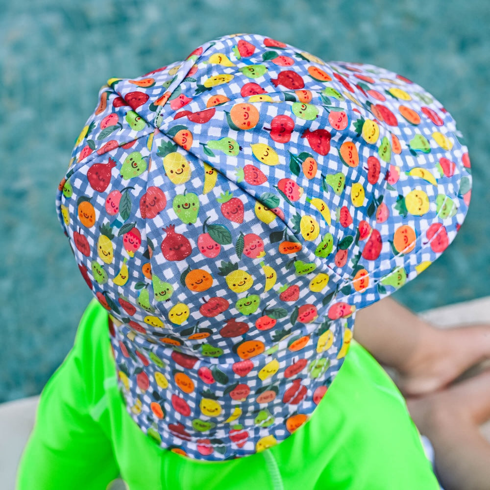 A back view of a child wearing a swim hat  featuring a blue and white gingham pattern and a colourful print of smiling cartoon fruits, including pineapples, strawberries and oranges. 