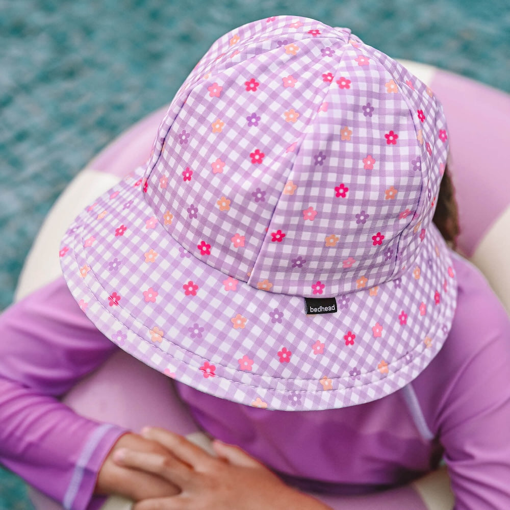 A child wearing a lavender and white gingham swim bucket hat with small pink, purple, and orange flowers, paired with a matching purple long-sleeve rashie, sitting in a pink and white pool float.