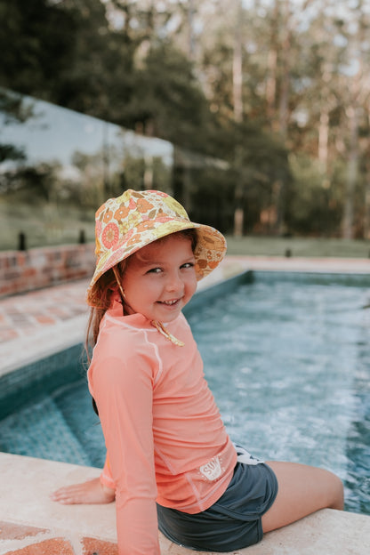 front view of Bedhead Tallulah Swim Bucket Hat worn by a young model sitting on the edge of a swimming pool