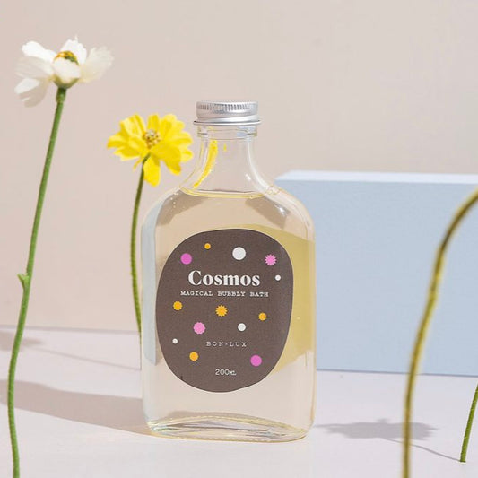 Bon Lux Cosmos Magical Bubbly Bath in a glass bottle, styled with yellow and white flowers on a white background.