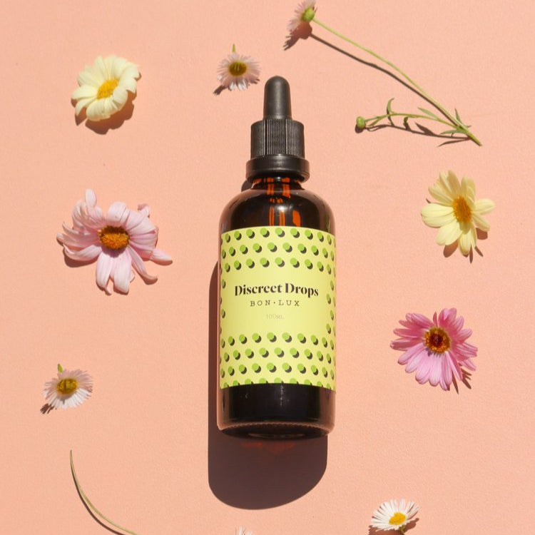 Bottle of Bon Lux Discreet Drops with a yellow label on a pink background with flowers.
