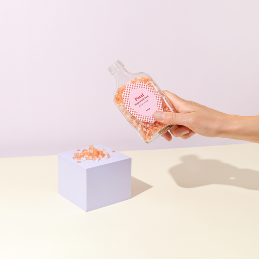 Hand holding a bottle of Bon Lux Petal Magical Salt Soak containing pink bath salts, with a small plinth with bath salts sprinkled on top.