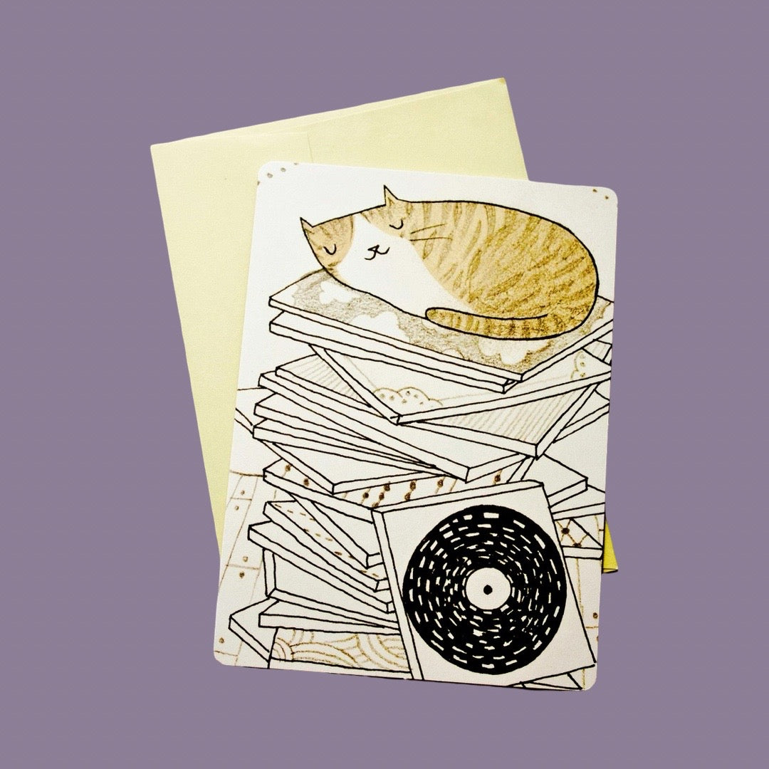 A greeting card with an illustration of a sleeping orange-and-white cat resting on a stack of vinyl records. A pale yellow envelope is placed behind the card against a solid lavender background.