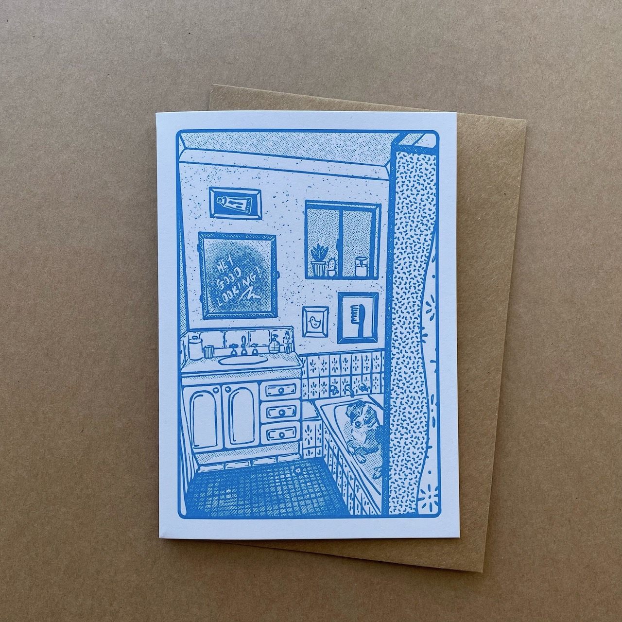 A greeting card featuring a blue line-art illustration of a bathroom interior with the mirror above the sink reading "hey good looking" and a border collie in the bathtub, placed on top of a brown kraft envelope against a brown kraft paper background.