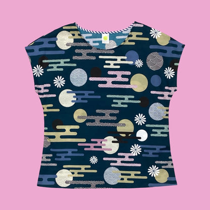 Essaye Tille short-sleeved top with a dark teal base, featuring a colourful abstract pattern of circles, wavy lines, and floral motifs in pastel tones, laid flat against a soft pink background.