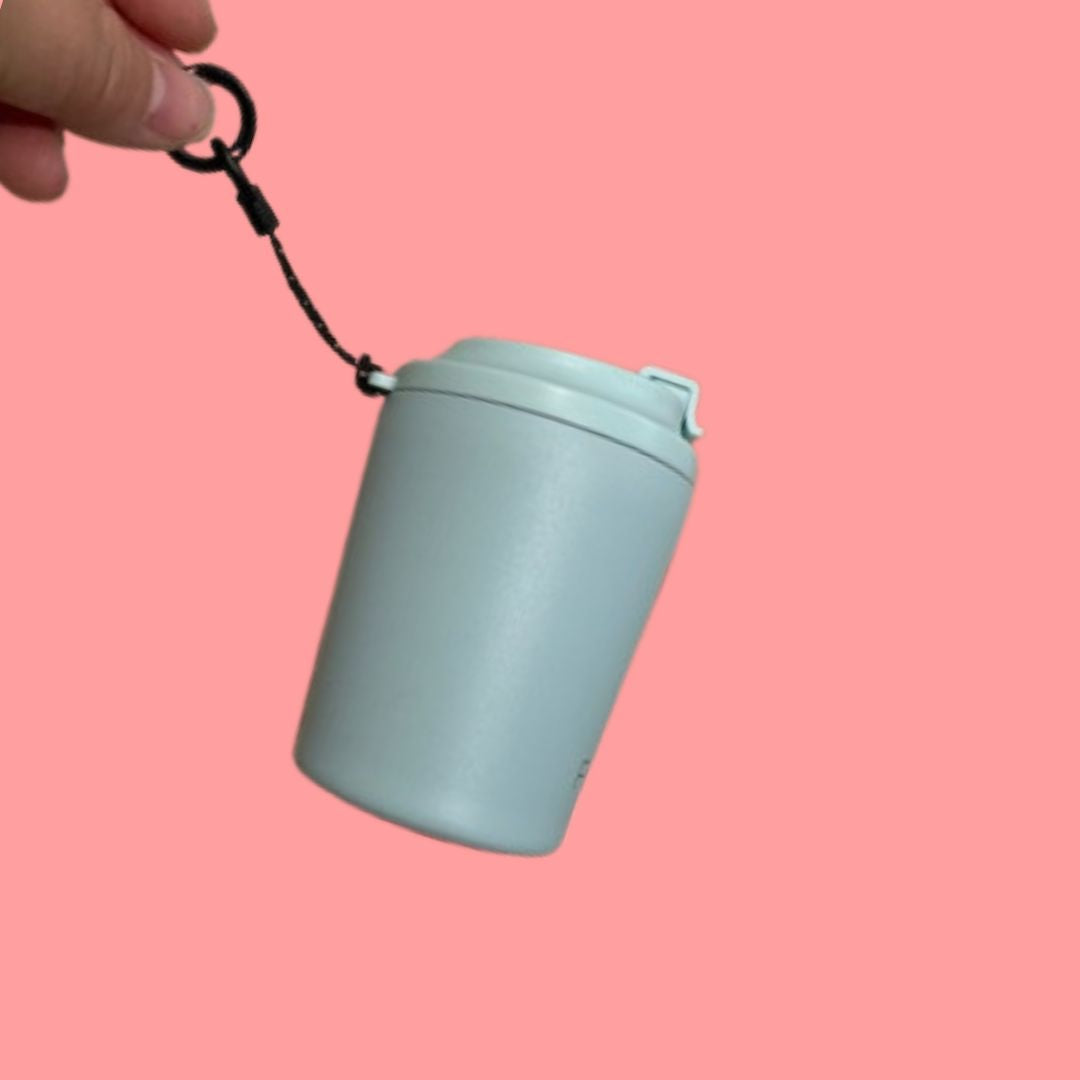 A hand is holding the hang loop of the Fressko piccolo reusable cup (a Small pale grey blue cup with a black strap) on a pink background