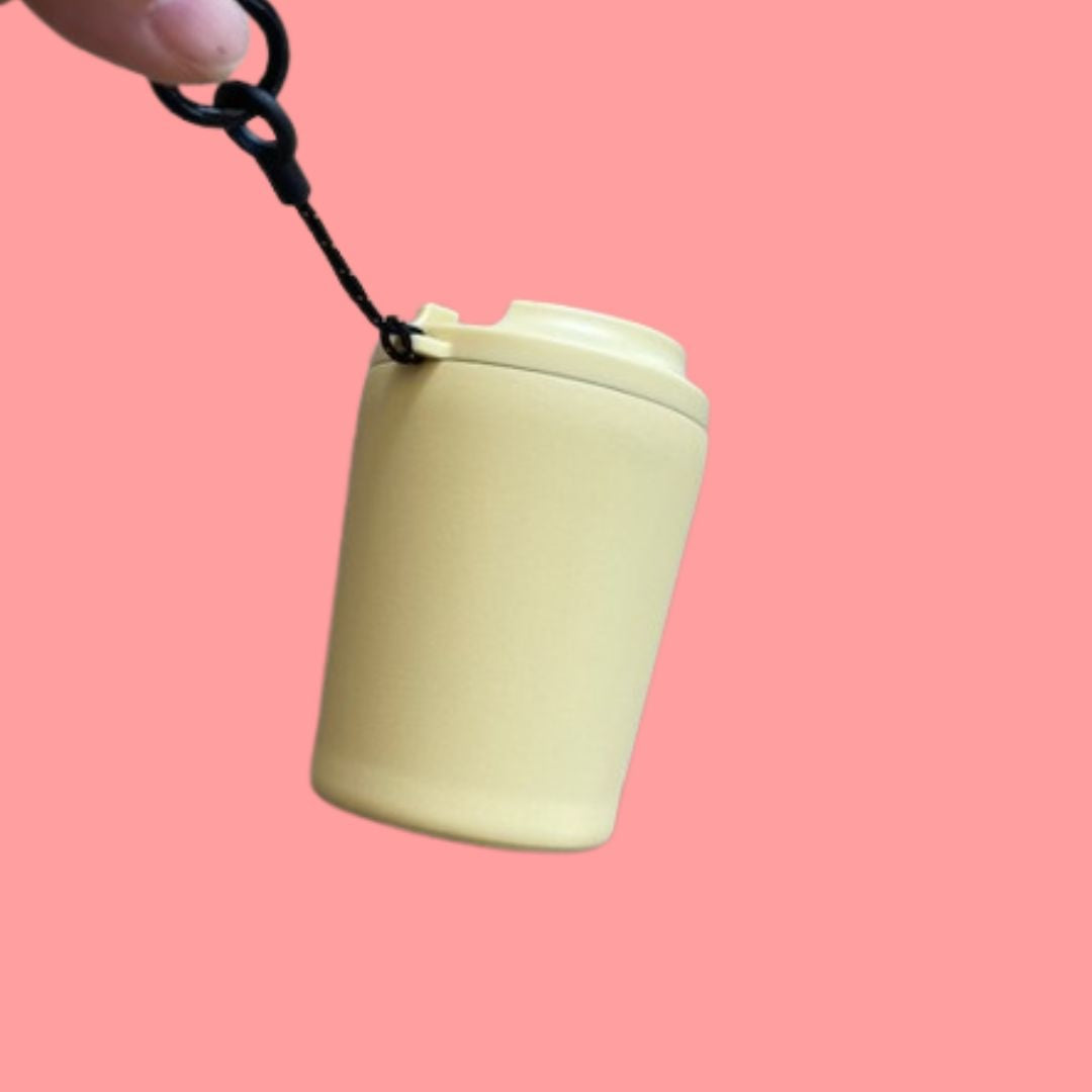 A hand is holding the black handle of the Light yellow travel mug on a pink background