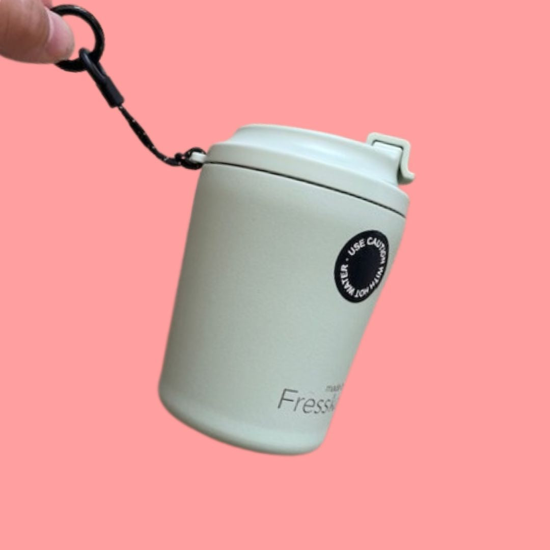 A hand is holding the black hang loop of the pale green reusable cup on a pink background