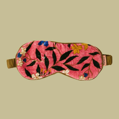 A pink velvet sleep mask with an embroidered floral and leaf pattern in black, yellow, blue, and white, featuring a brown elastic strap. The mask is displayed flat against a solid beige background.