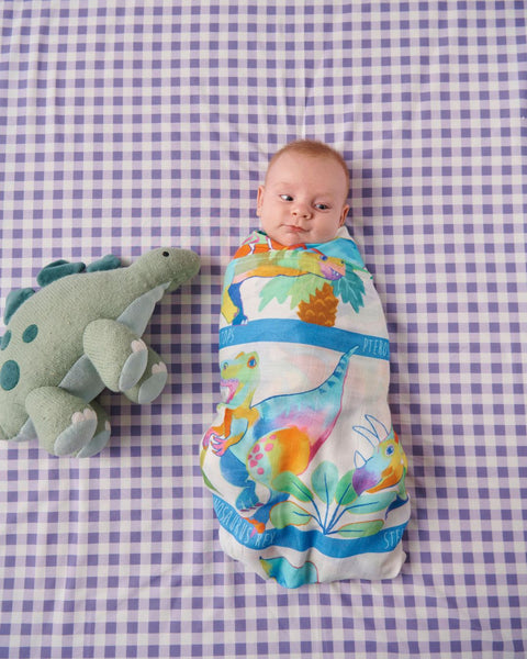 A bird's eye view of a baby lying in a bag wrapped up in a Kip & Co Dino Safari Bamboo Baby Swaddle. The bedsheet is a purple gingham pattern. And a stuffed toy dinosaur is lying next to the baby.