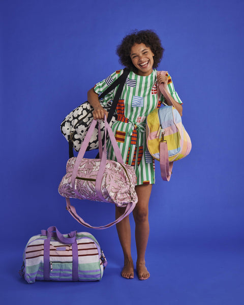 Model is posing with multiple Kip & Co duffle bags. She is holding the Kip & Co Palmy Duffle Bag, on her shoulders is a Kip & Co Corfu Tartan Duffle Bag, and on her other shoulder is a black and white duffle bag. On the floor next to her is the Kip & Co Island Delight Duffle Bag.