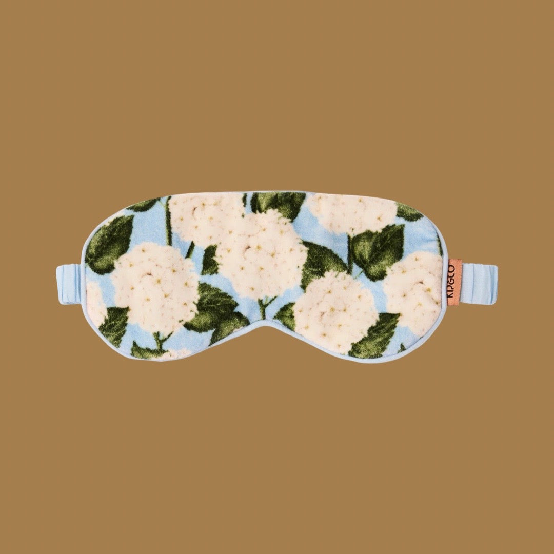 A plush sleep mask with a light blue background and a white flower and green leaf pattern, featuring light blue straps and a small orange brand tag that reads 'kip&co'. Background is a solid light brown.