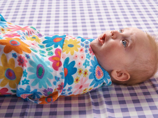 Baby lying in a crib wrapped in a Kip & Co Rainbow Flowers Bamboo Baby Swaddle.