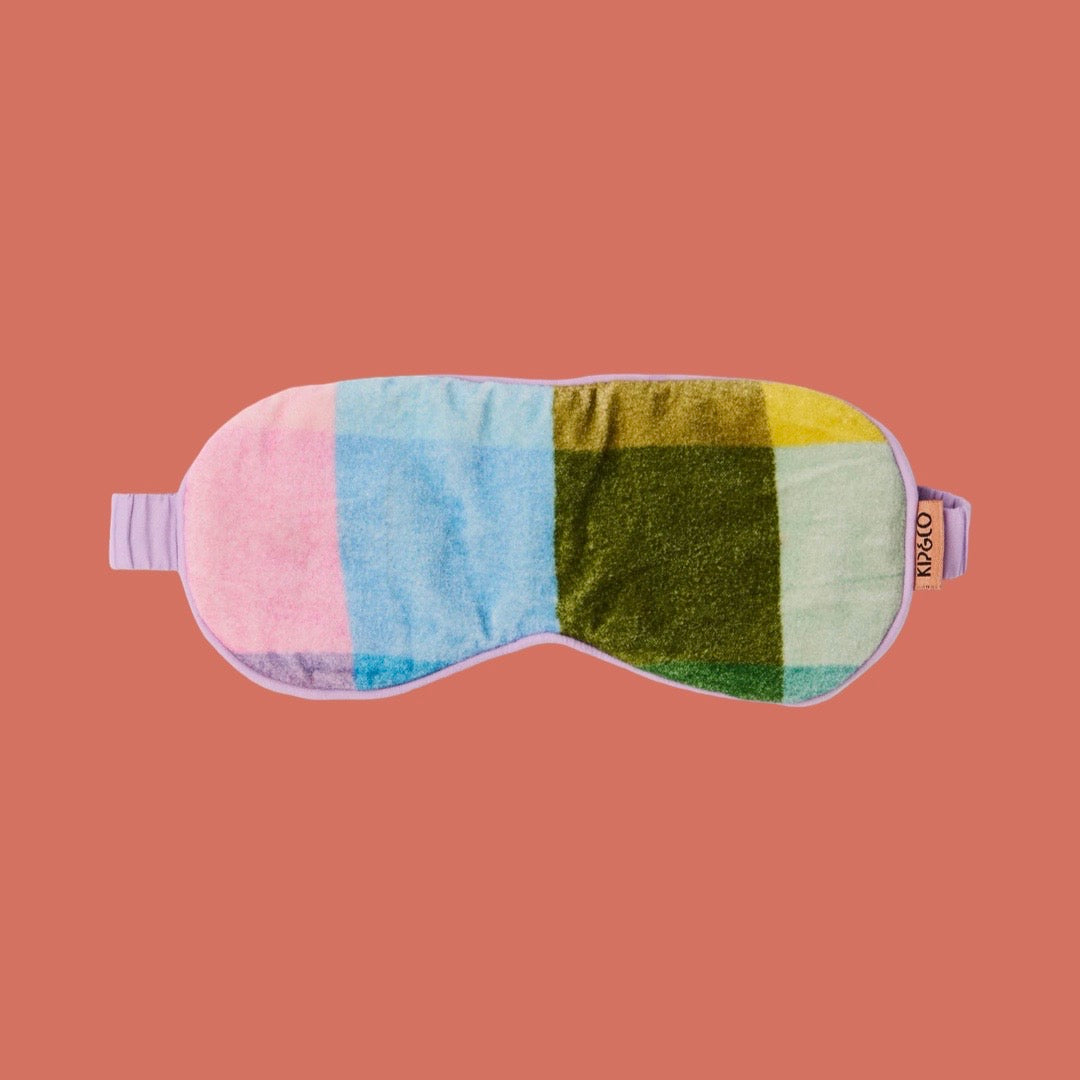 A plush eye mask with a colorful patchwork design in pink, light blue, olive green, and yellow, featuring a lavender elastic strap and a small brand tag that reads 'kip&co'. The mask is centred on a solid terracotta background.