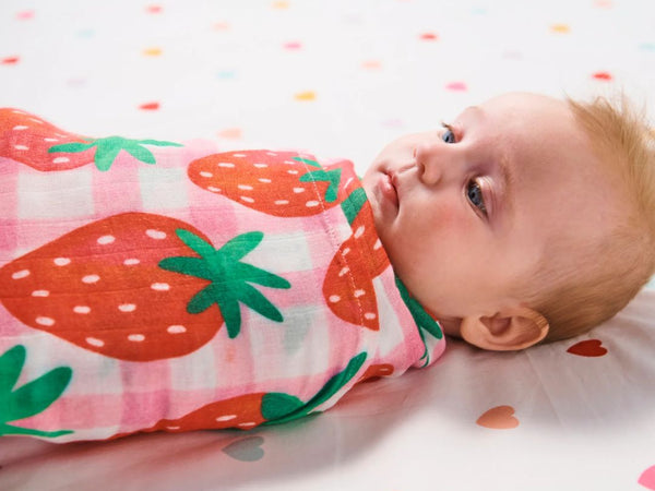 Landscape view of a baby lying in a bed wrapped in a Kip & Co Strawberry Jam Bamboo Baby Swaddle.