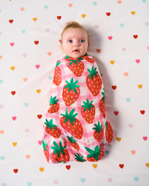 A bird's eye view of a baby lying in a bed wrapped up in a Kip & Co Strawberry Jam Bamboo Baby Swaddle. The bedsheet is covered with multicoloured love hearts.