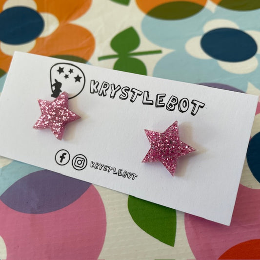 Krystlebot Star Studs - Pink Glitter. Pink star-shaped earrings on a KRYSTLEBOT branded card with a colorful floral background.