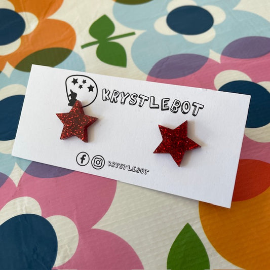 Krystlebot Star Studs - Red Glitter. Red star-shaped earrings on a 'KRYSTLEBOT' branded card with a colorful floral background.