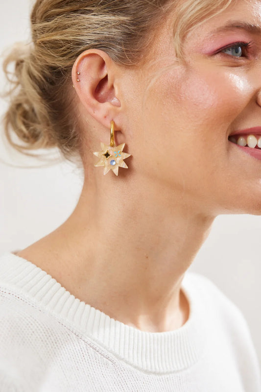 A close-up of a woman wearing a gold starburst earring with two small stones in the centre against a soft light background.