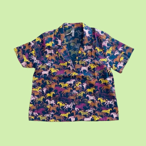 Mata Traders Camp Shirt in Horses. This collared button-down shirt features a pattern full of horses in different colours with a dark blue background.