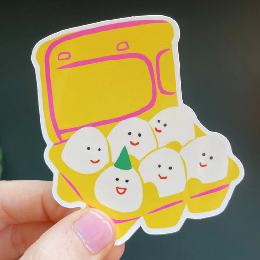 Sticker by Naomi Wilkinson of a yellow egg carton with six smiling eggs, held by a hand.