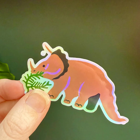 Holographic triceratops sticker by Naomi Wilkinson, held by a hand on a green background