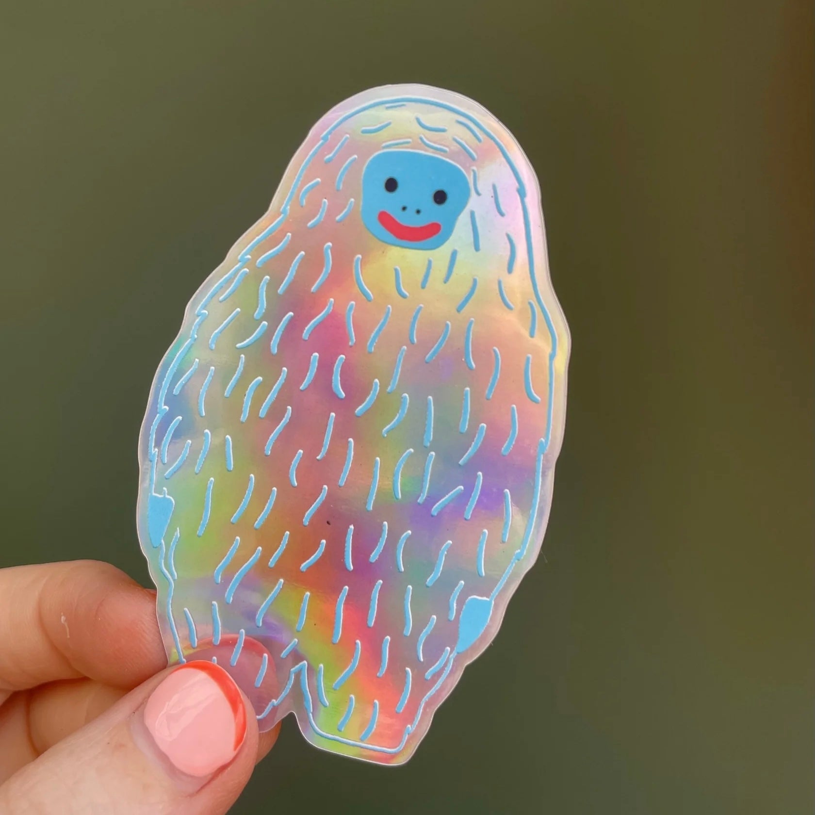Holographic sticker by Naomi Wilkinson of a smiling yeti with a blue face, held by a hand against a dark background