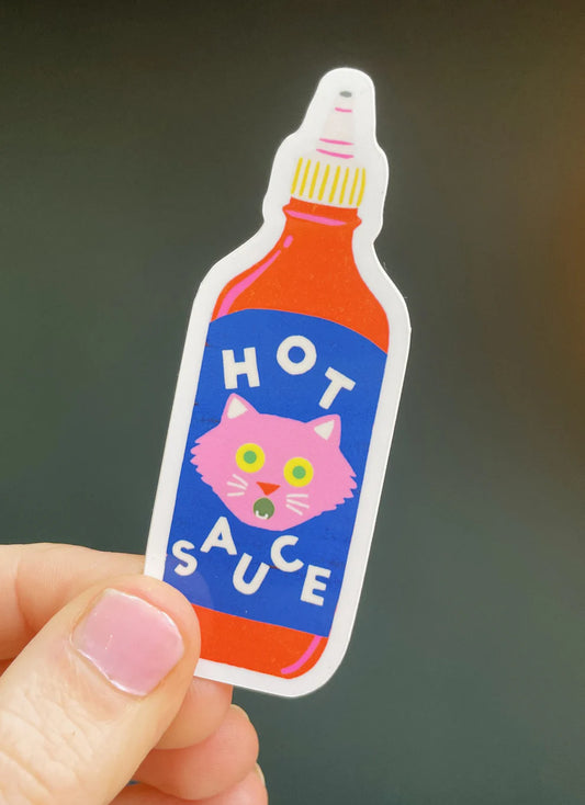 Sticker by Naomi Wilkinson of a hot sauce bottle with a cat face, held by a hand against a dark background
