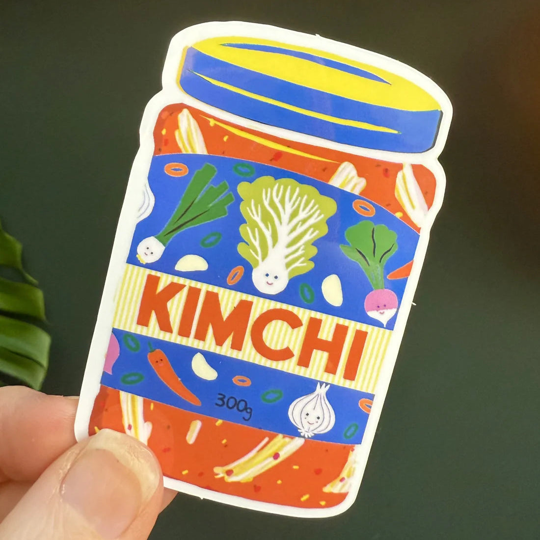 Colorful sticker of a kimchi jar held by a hand against a dark background