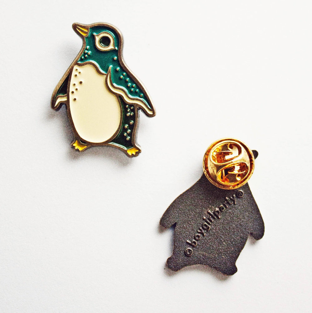 Two penguin-shaped pins on a white background. One pin shows a teal, cream, and yellow penguin, and the other shows the back in dark gray with a gold pin backing.