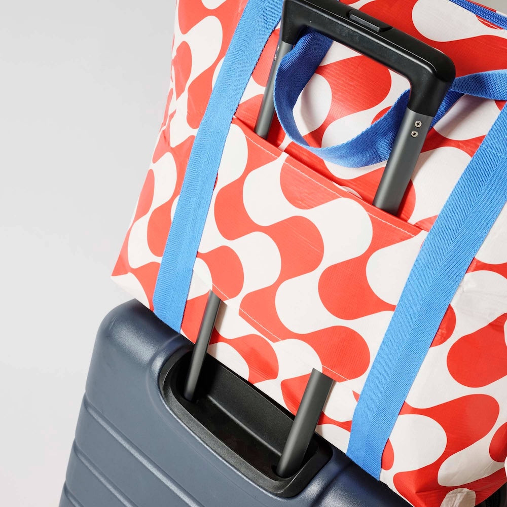 A close-up of a Project Ten Red Ripple Medium Zip Up Tote with blue handles, shown resting on top of a dark grey rolling suitcase with the suitcase handle through the tote's sleeve, against a white background.