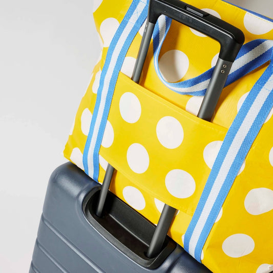 Styled image of a close up of Project Ten Yellow Polka Dot Medium Zip Up Tote showing off the sleeve feature draped over a dark grey rolling suitcase against a white background.