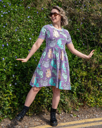 Woman wearing a purple dress with cat illustration print standing in front of green foliage.