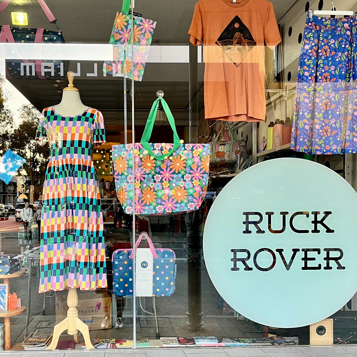 Ruck Rover's window display featuring colourful patterned clothing and bags. A mannequin wears a short-sleeve dress with a multicolour geometric stripe pattern, a large tote bag with a floral daisy print and green handles hangs in the centre, and a blue polka-dot laptop is displayed below.