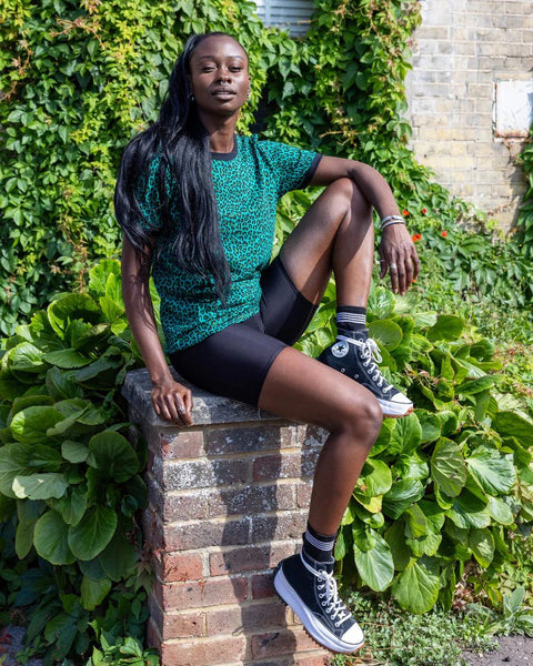 Model is wearing Run and Fly Leopard Tee Shirt in Teal and paired with black shorts. She is sitting on a brick bollard, and facing the camera.