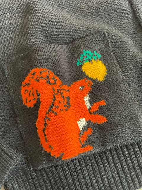 Close-up of a black knit sweater featuring a knitted pocket with an orange squirrel and a yellow acorn with green leaves.